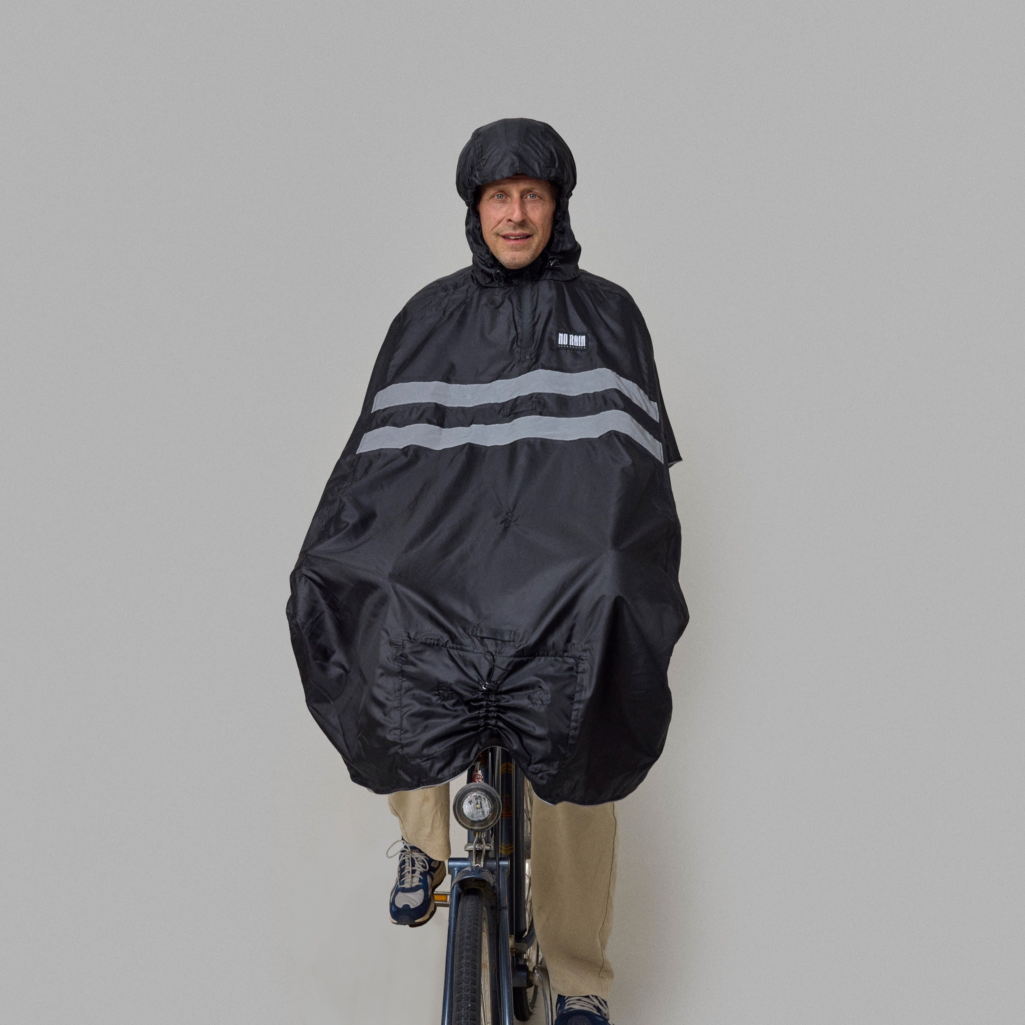 Person wearing a black NoRain bicycle poncho with reflective stripes while riding a bicycle on a gray background