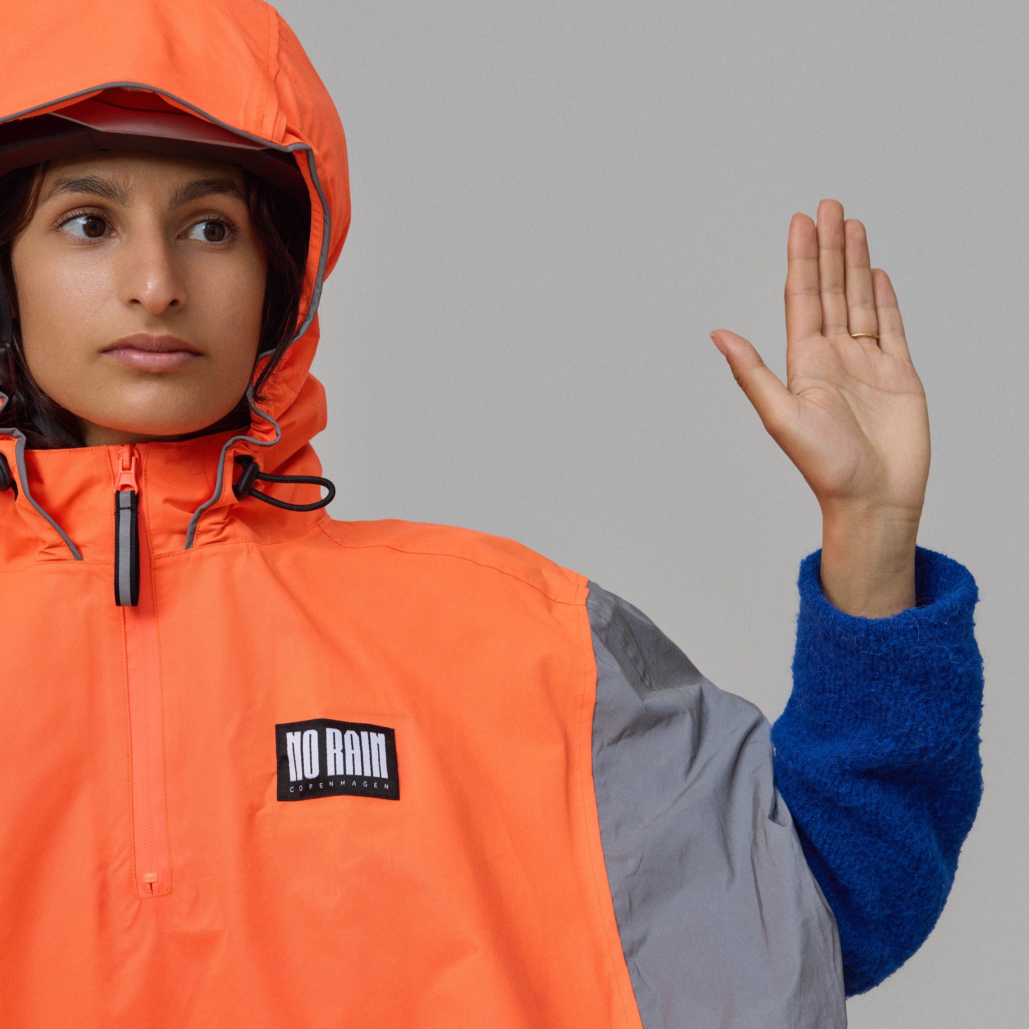 Person wearing an orange and gray NoRain bicycle poncho with a brand logo, raising their hand against a gray background