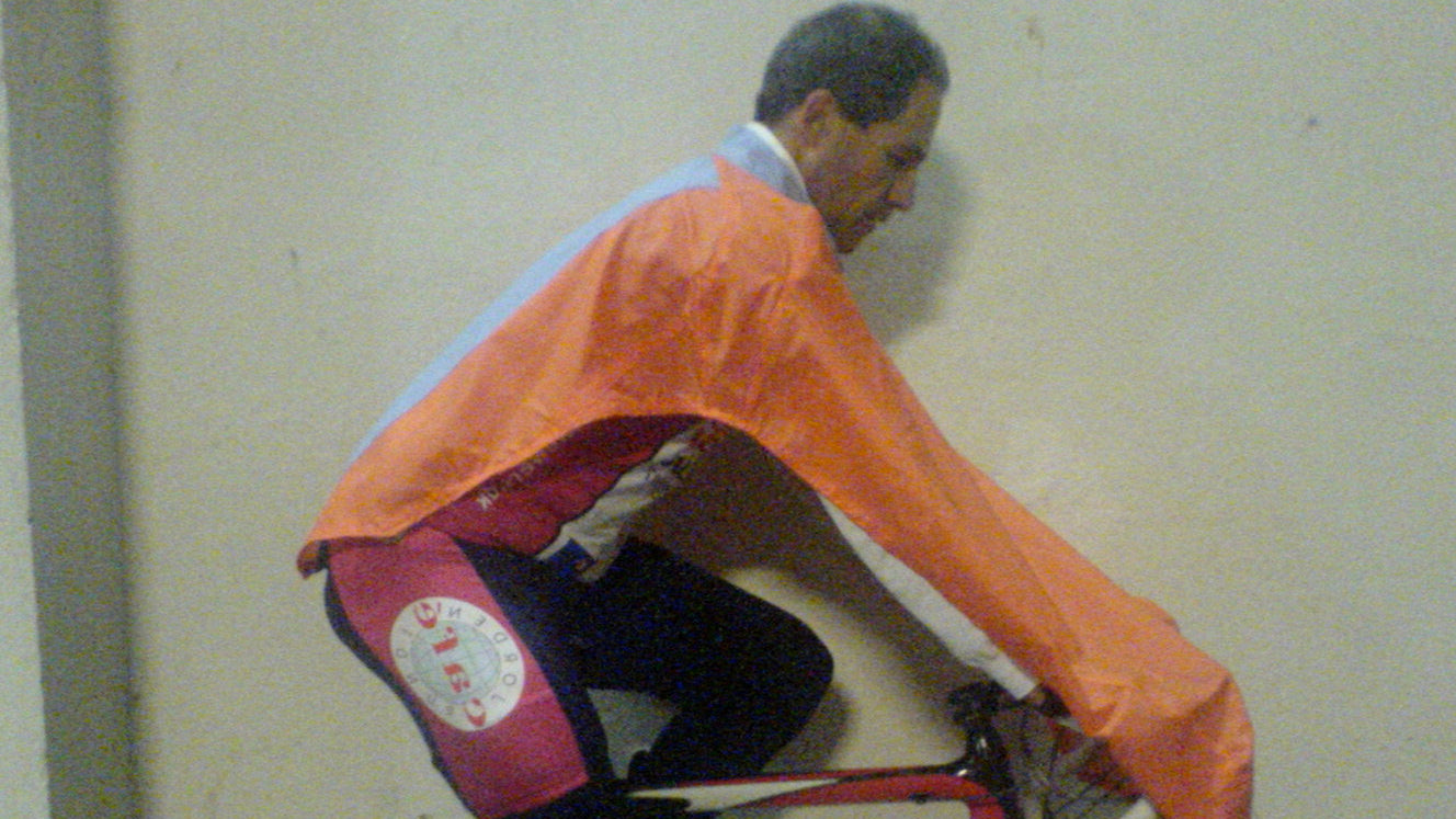 Person on a bicycle wearing an orange NoRain Bicycle Poncho against a plain wall.