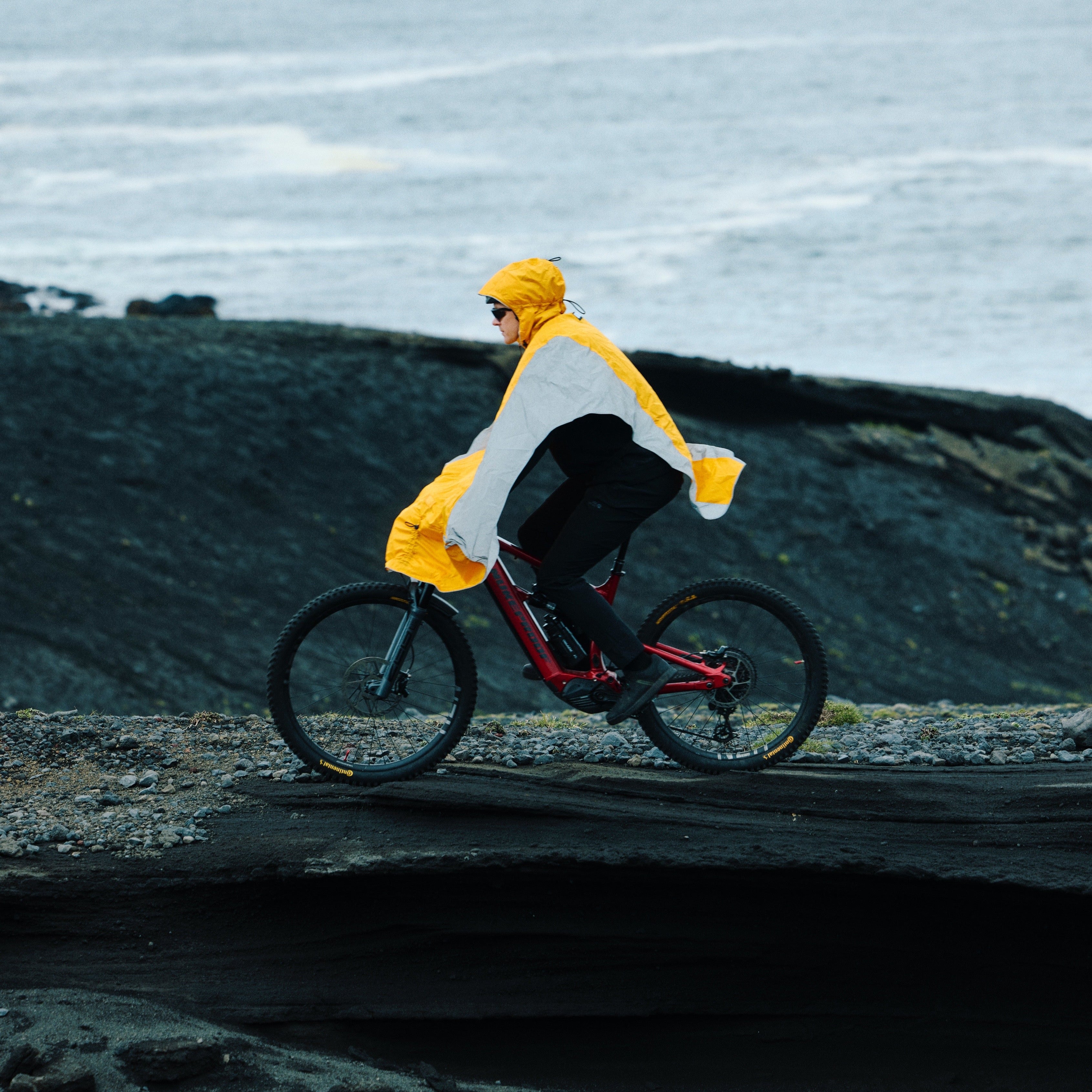 Person riding a bicycle with a NoRain Copenhagen Poncho on a rocky coastal path with a yellow raincoat.