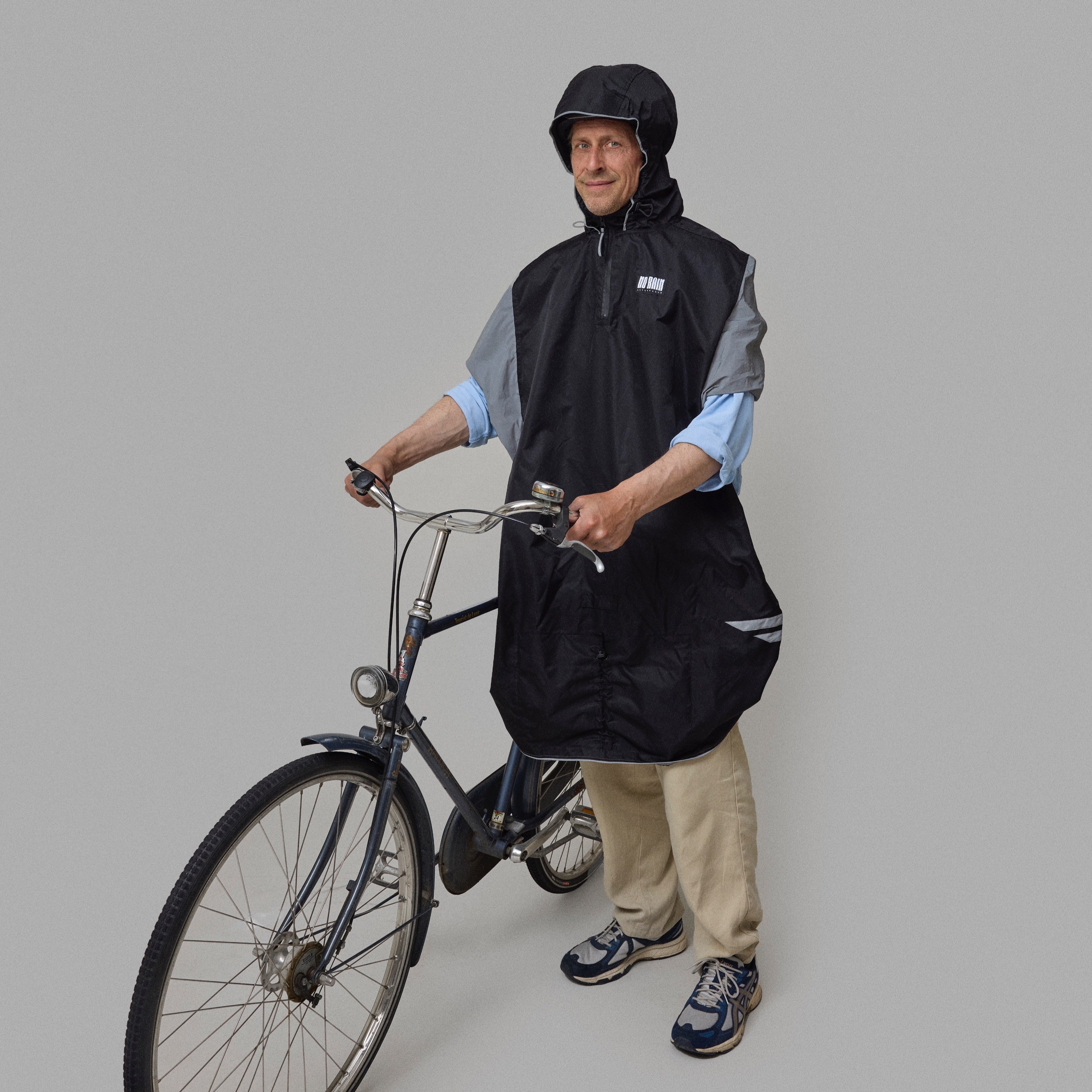 Person wearing a black NoRain Bicycle poncho and helmet, standing next to a bicycle on a gray background