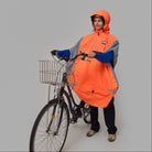 Person wearing an orange NoRain bicycle poncho standing next to a bicycle on a gray background