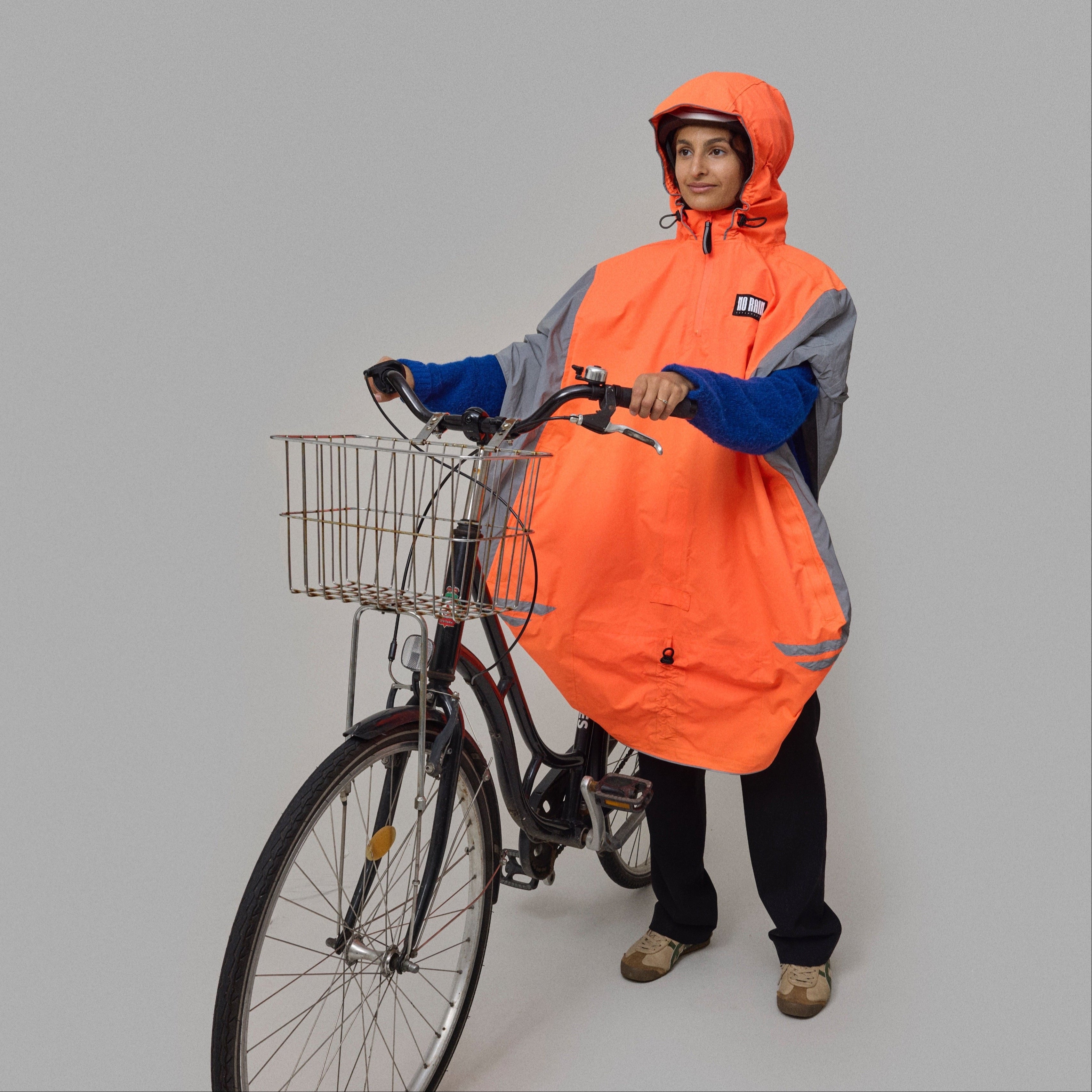 Person wearing an orange NoRain bicycle poncho standing next to a bicycle on a gray background