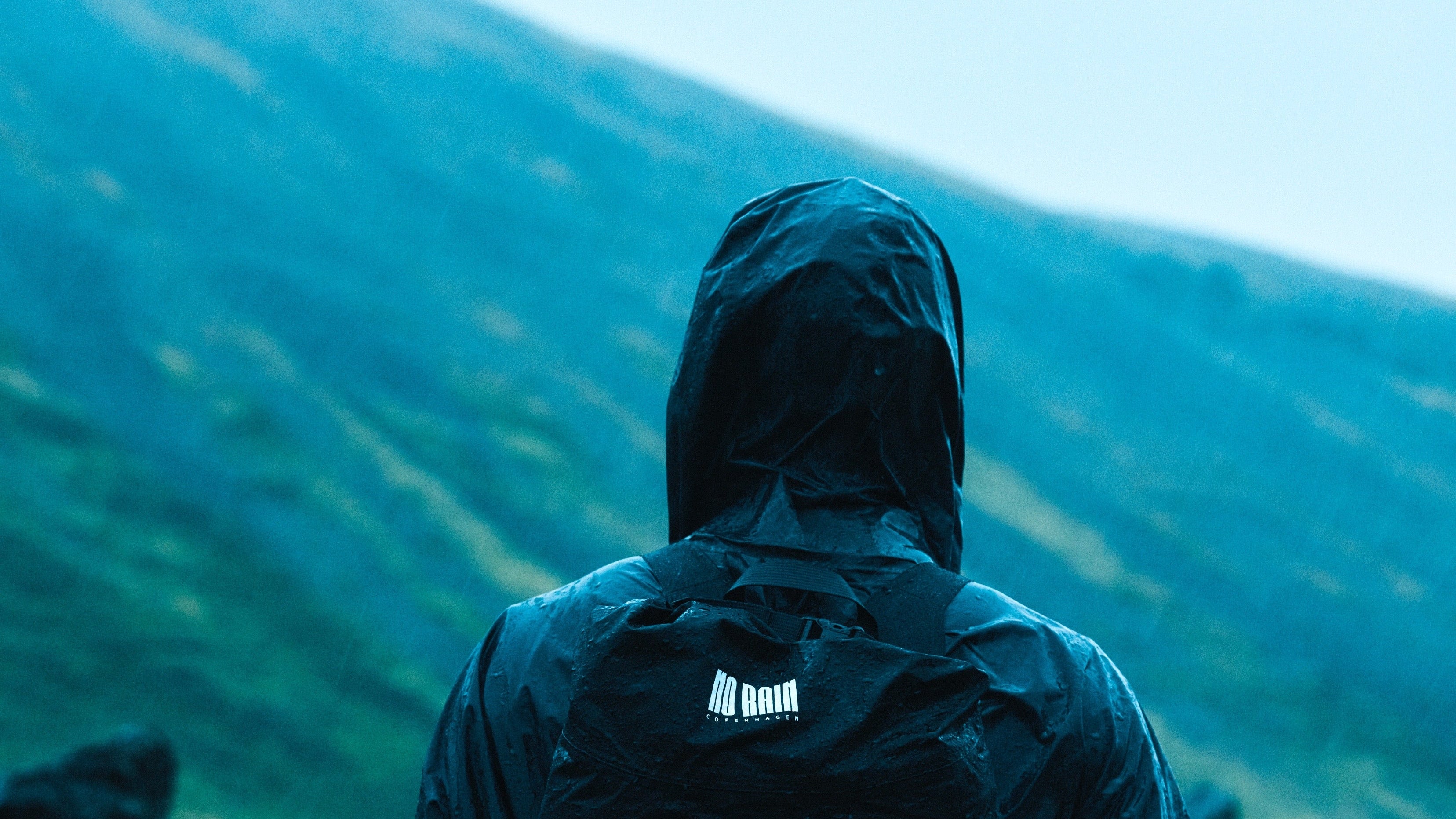 Person wearing a dark jacket with a NoRain logo standing on a mountainous landscape.
