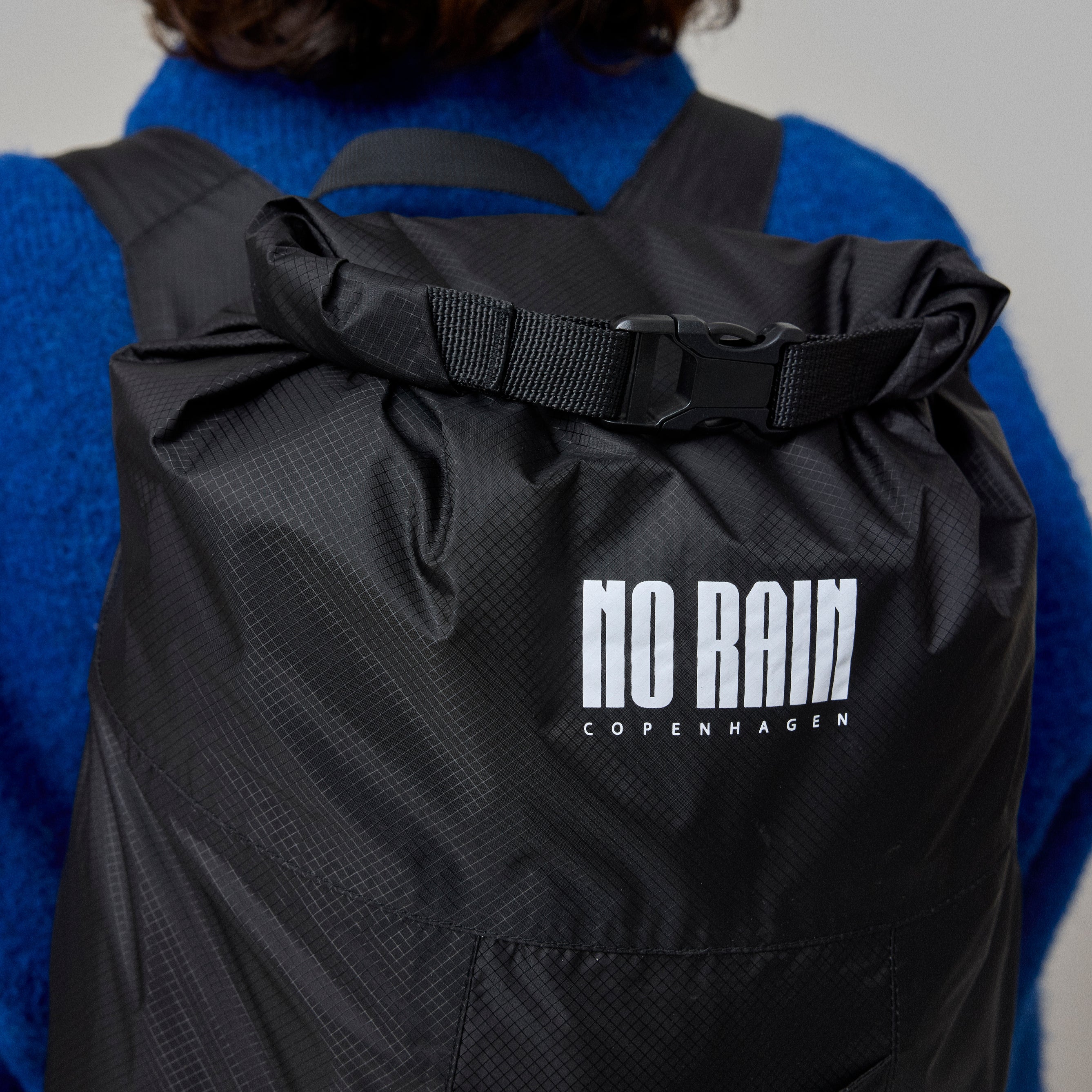 Person wearing a black waterproof backpack with 'NO RAIN Copenhagen' logo on a plain background