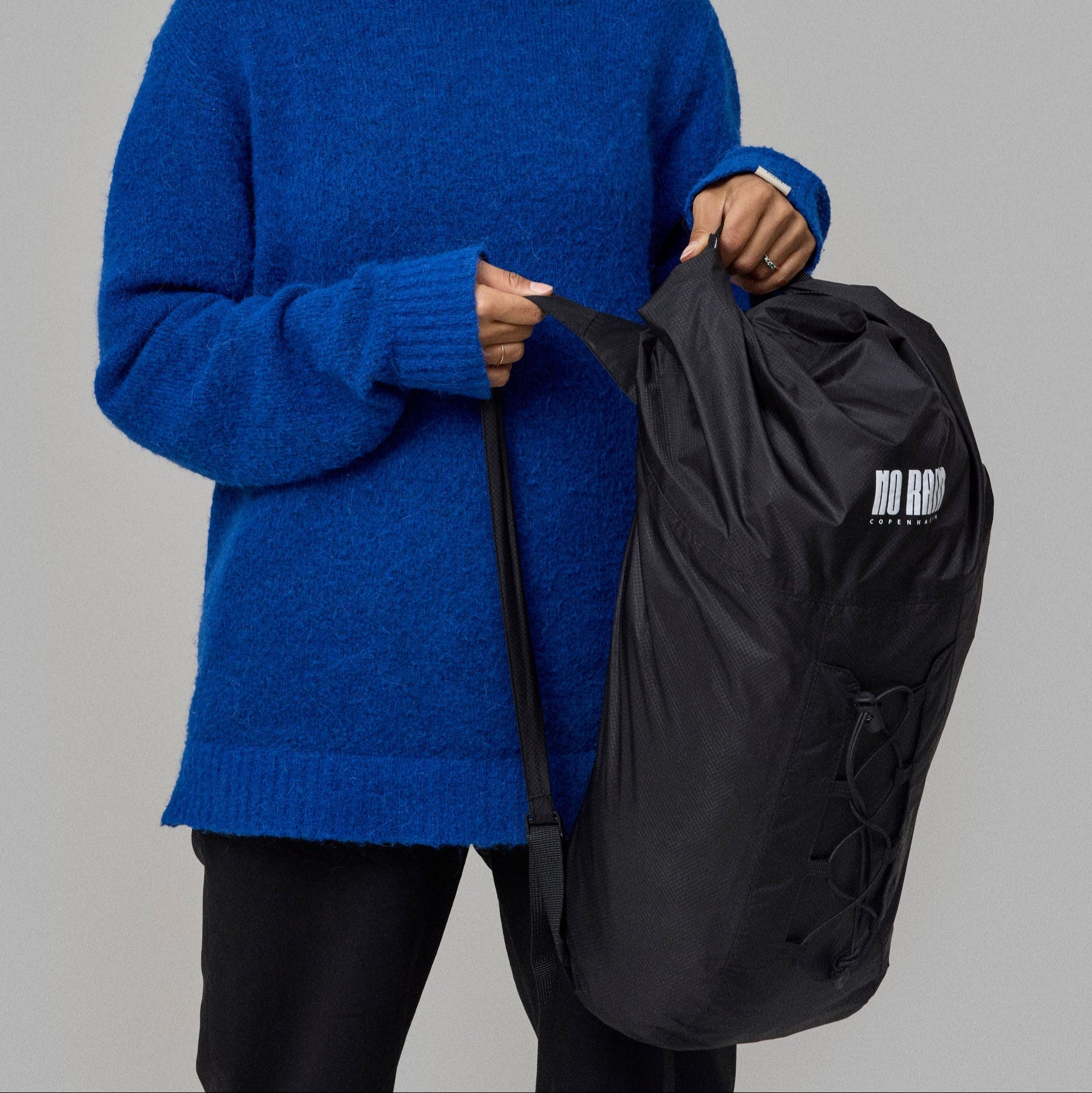 Person wearing a blue sweater holding a black waterproof norain backpack against a gray background